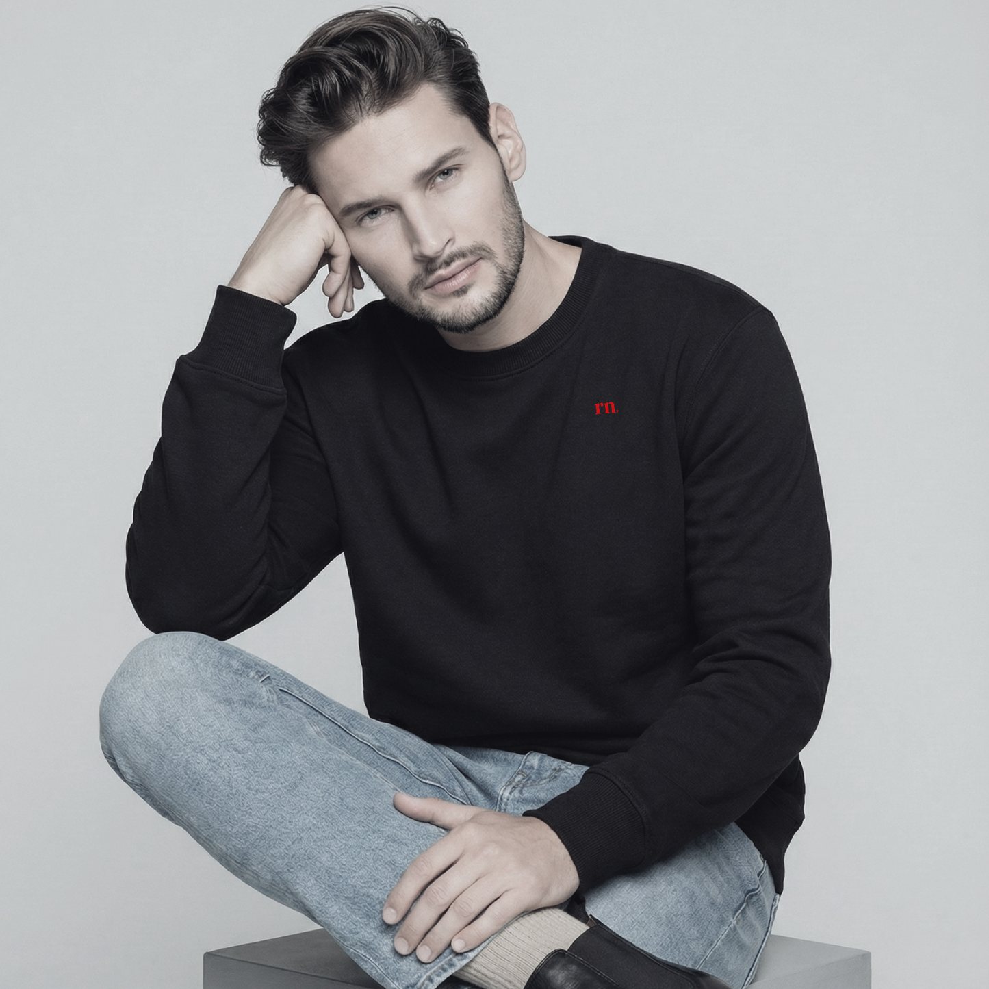 Male model wearing the Rascal Nation black crewneck sweatshirt with red rn. logo, paired with jeans and black boots in a clean studio setup.